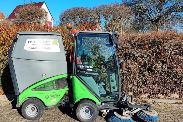 Grüner Kehrmaschine neben einer Hecke vor einem hellen Himmel.