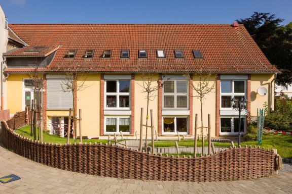 Beige Gebäude mit rotem Dach, großen Fenstern und Spielbereich im Freien.