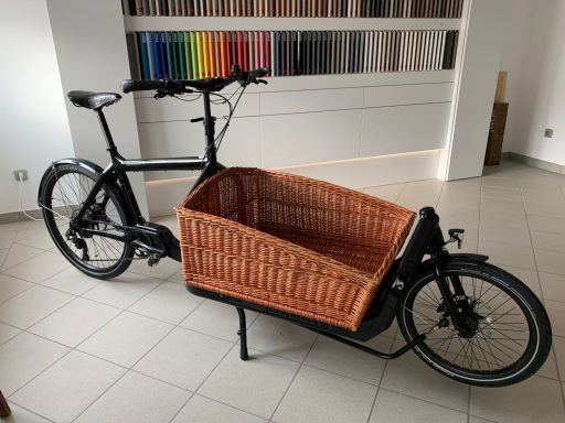 Schwarzes Lastenfahrrad mit Korbanhänger aus Rattan in einem hellen Raum.