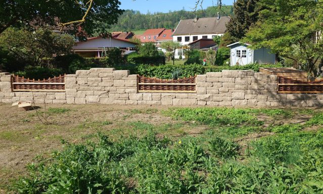 Eine Mauer aus Steinen, umgeben von Gras und Bäumen, im Hintergrund ein Wohngebiet.