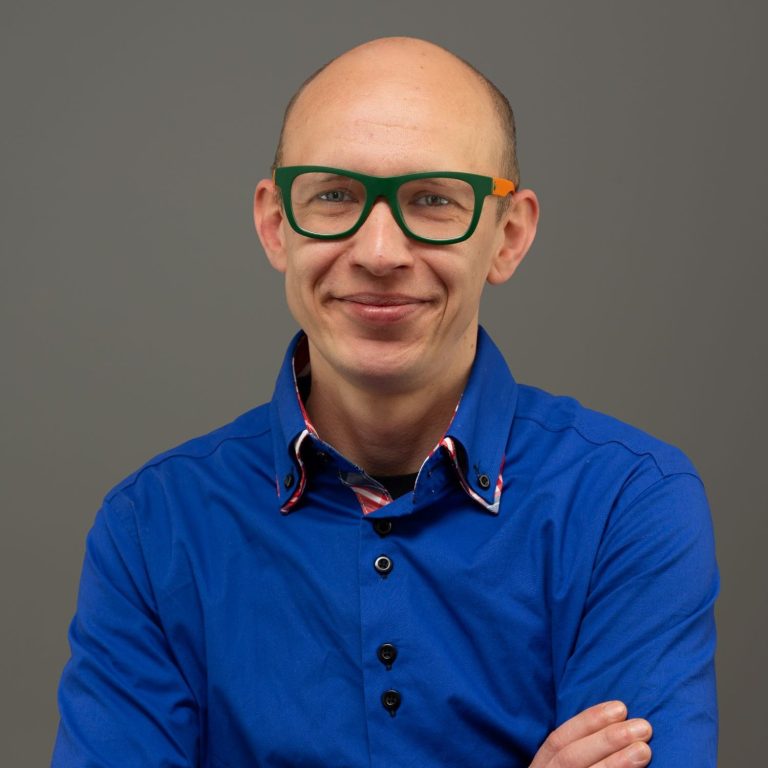 Mann mit Glatze und grünen Brillen, trägt ein blaues Hemd mit buntem Kragen.