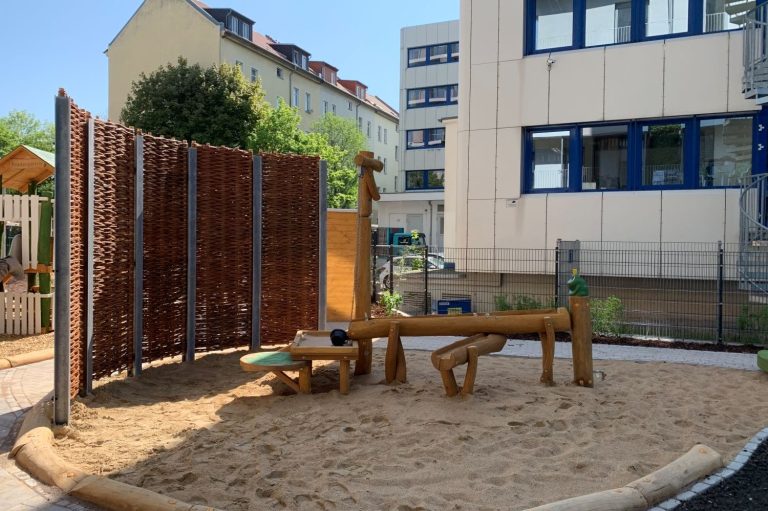 Spielplatz mit Sandfläche, Holzspielgeräten und Sichtschutzwänden.