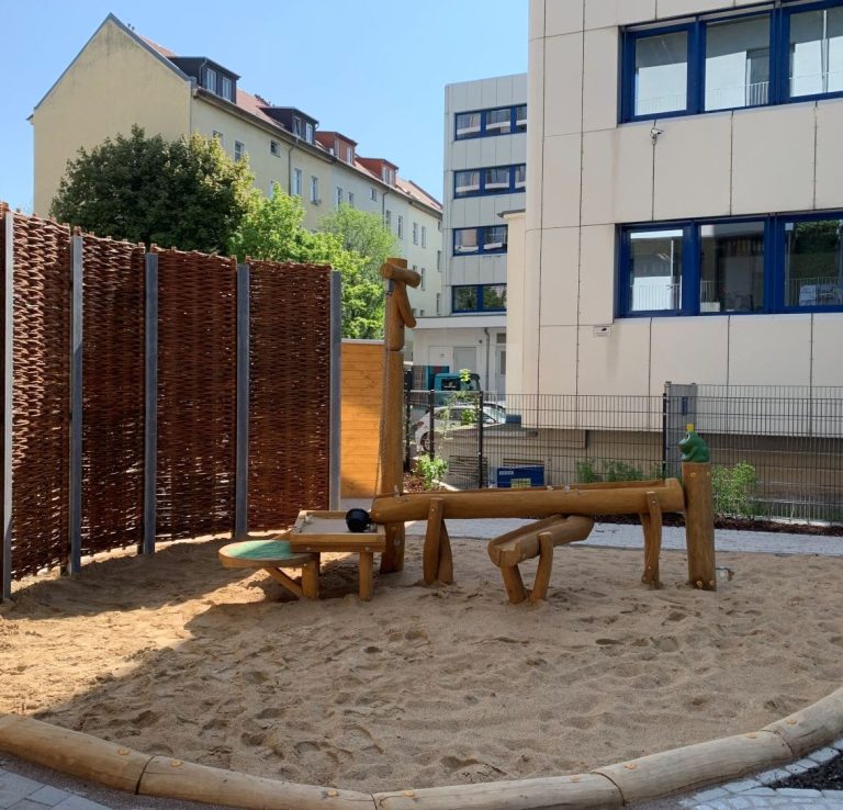 Sandspielplatz mit Holzspielgeräten und Umrandung aus Flechtzaun.