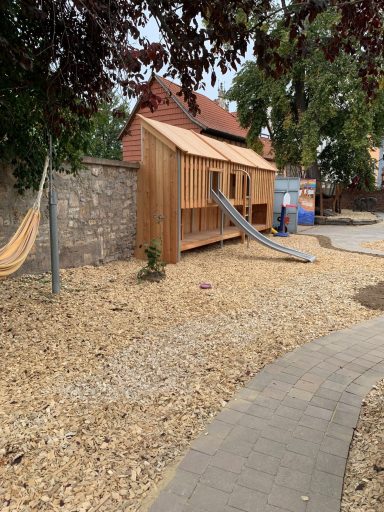 Holzhütte mit Rutsche und Sandfläche in einem Spielplatzbereich.