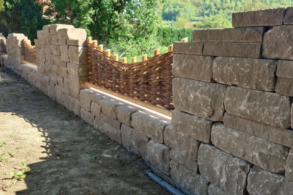 Steinmauer mit gebogenem Holzgittern zwischen den Steinen, umgeben von Natur.