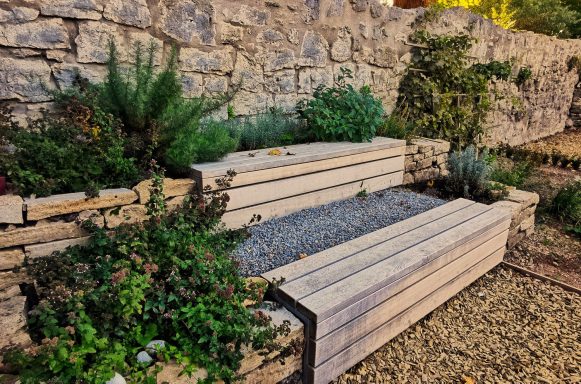 Holzterrasse mit Pflanzen in einem gepflegten Garten, Steinwand im Hintergrund.