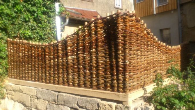 Holzlattenzaun mit einer gewellten Oberkante, umgeben von Pflanzen und einem Gebäude im Hintergrund.