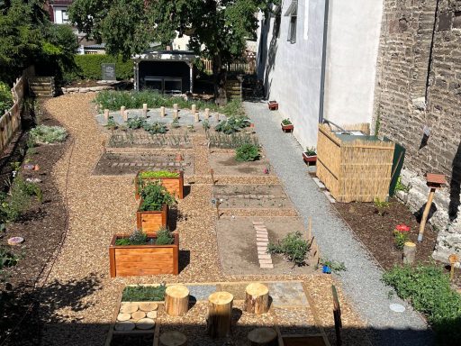 Gemüsegarten mit Hochbeeten, Wegen aus Kies und Holzstühlen, umgeben von Pflanzen.
