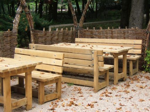 Holzbank und Holztische im Freien, umgeben von Bäumen und Laub.