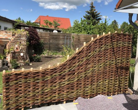 Ein geschwungener Zaun aus geflochtenem Material in einem Garten mit Pflanzen und Bäumen.