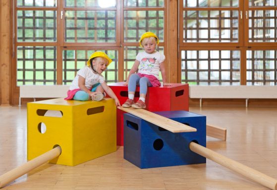 Zwei Kinder auf bunten Kisten mit Helmen, spielen und lachen in einem hellen Raum.