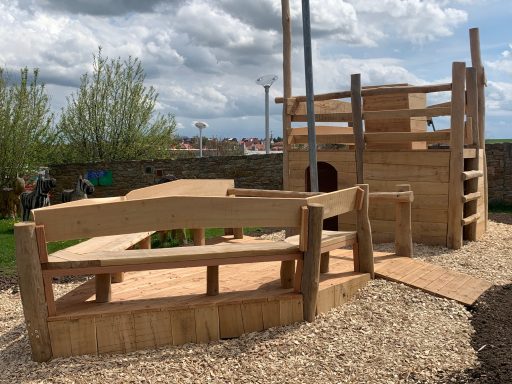 Spielplatz aus Holz mit Kletterstruktur und Sitzgelegenheiten, umgeben von Rindenmulch.