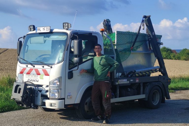 Mann steht neben einem kleinen Lkw mit einem Ladecontainer auf einem Feldweg.