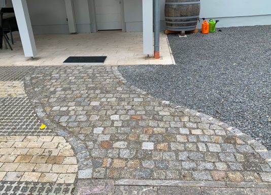 Steinweg mit geschwungenem Muster, umgeben von Schotter und einem grauen Gebäude.