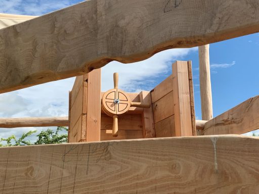 Holzapparat mit einem Rad, umgeben von Holzbalken und grünen Pflanzen im Hintergrund.
