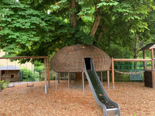 Spielplatz mit einem Baumhaus und einer Rutsche, umgeben von Bäumen und Spielgeräten.