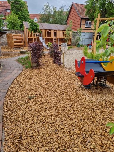 Spielplatz mit Holzspielsachen, Kiesweg und bunten Pflanzen im Garten.