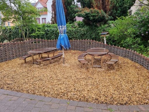 Spielplatz mit Holzstühlen und Tischen, umgeben von Rindenmulch und Pflanzen.