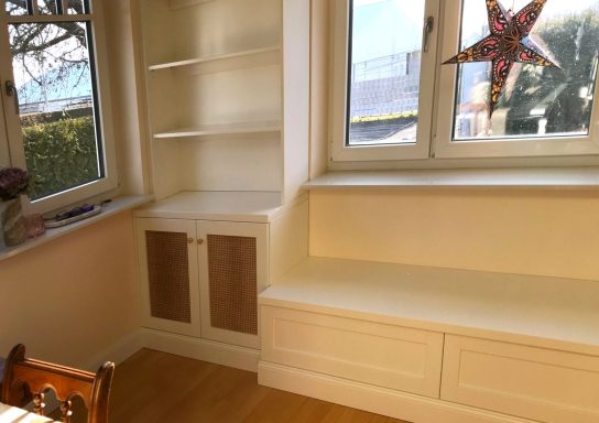 Ein heller Raum mit einem Fensterplatz, Bücherregal und Schrank in neutralen Farben.